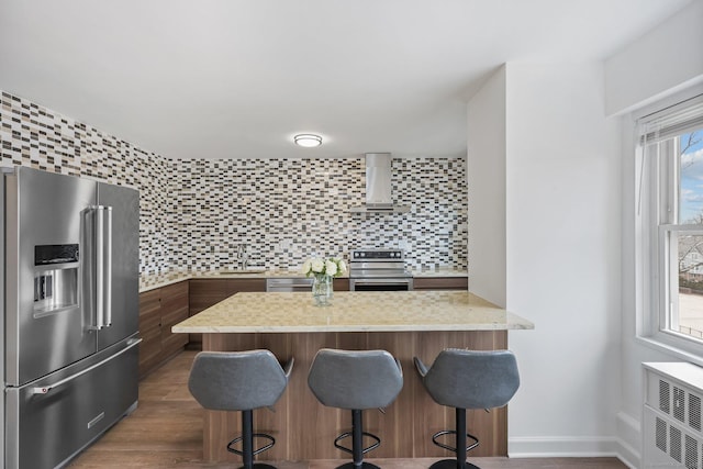 kitchen with stainless steel appliances, a peninsula, wall chimney range hood, decorative backsplash, and a kitchen bar