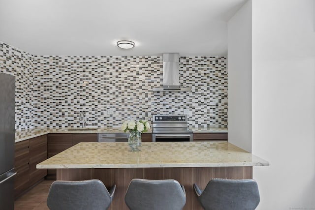 kitchen with a breakfast bar area, stainless steel appliances, decorative backsplash, a sink, and wall chimney exhaust hood