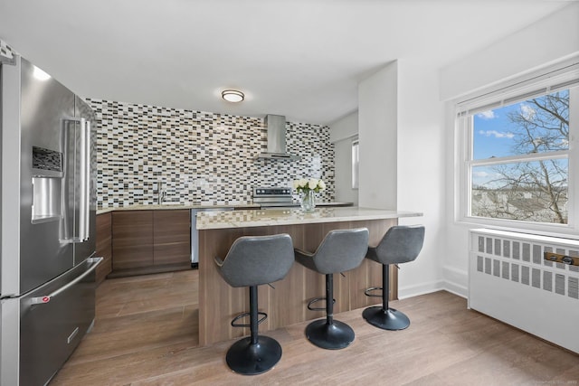 kitchen featuring stainless steel appliances, backsplash, radiator heating unit, wall chimney range hood, and modern cabinets