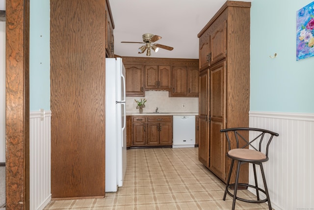 kitchen featuring ceiling fan, a wainscoted wall, light floors, brown cabinetry, and white appliances