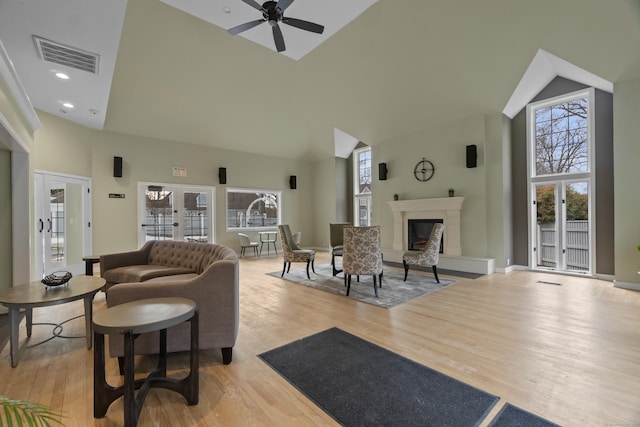 living area featuring light wood finished floors, visible vents, a glass covered fireplace, ceiling fan, and high vaulted ceiling