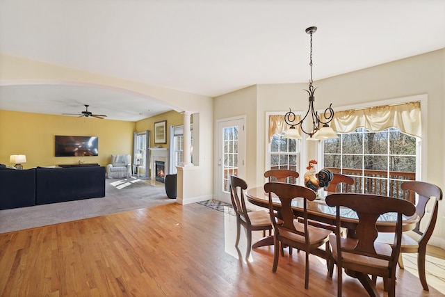 dining area featuring arched walkways, wood finished floors, a lit fireplace, ornate columns, and ceiling fan with notable chandelier