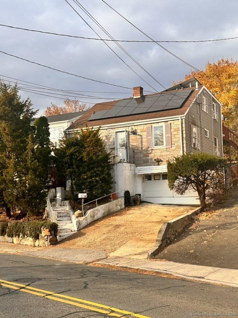 view of front of home featuring driveway, a garage, stone siding, a chimney, and roof mounted solar panels