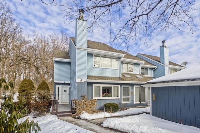 snow covered rear of property with a shingled roof and a chimney