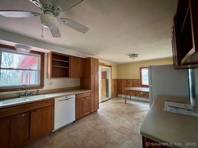 kitchen featuring white dishwasher, wood walls, a sink, wainscoting, and brown cabinetry