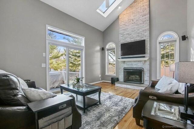 living room with high vaulted ceiling, wood finished floors, a skylight, baseboards, and a brick fireplace