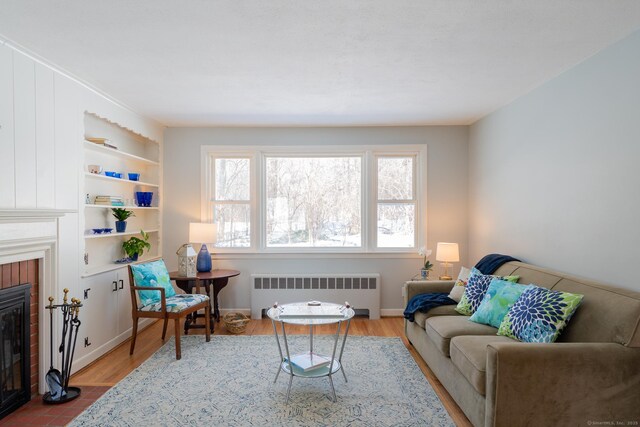 living area featuring built in shelves, wood finished floors, baseboards, a brick fireplace, and radiator heating unit