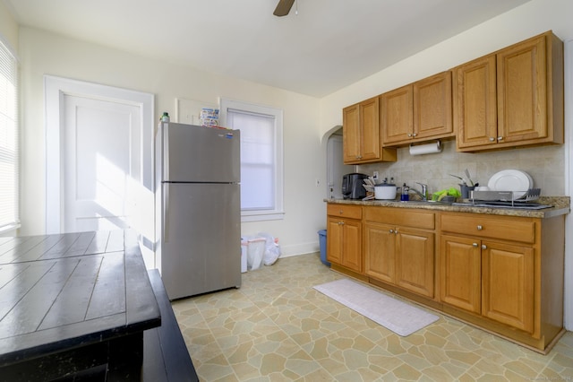 kitchen with arched walkways, brown cabinets, backsplash, a ceiling fan, and freestanding refrigerator