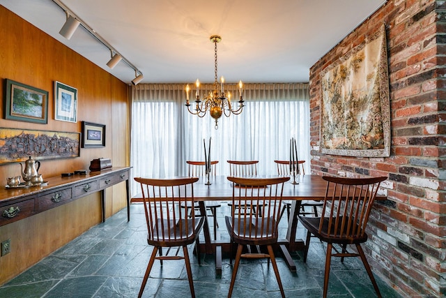 dining space with brick wall, stone finish floor, rail lighting, wood walls, and a notable chandelier