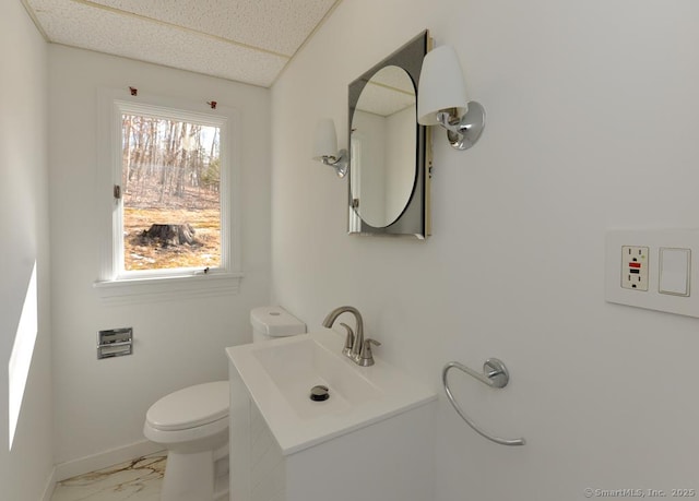 half bathroom with a drop ceiling, toilet, vanity, baseboards, and marble finish floor