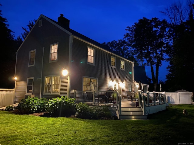 back of house at twilight with a lawn, a chimney, a wooden deck, and fence