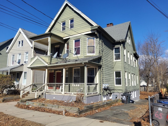 view of front facade featuring roof with shingles, a porch, a chimney, and a balcony