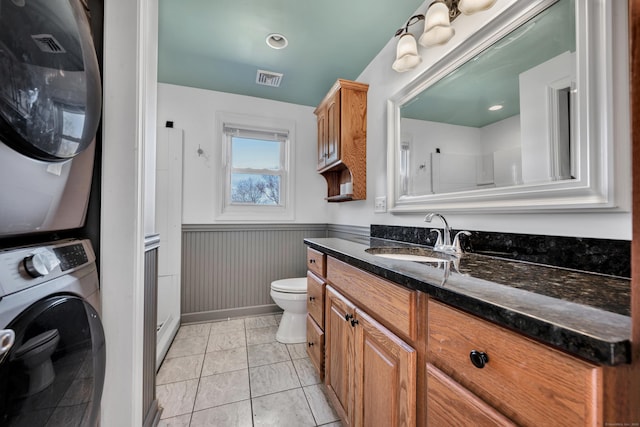 full bath featuring vanity, tile patterned flooring, stacked washer / drying machine, wainscoting, and toilet