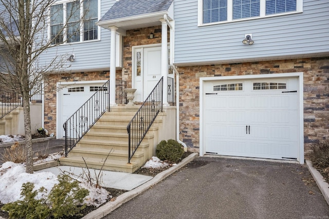 view of exterior entry with a garage, stone siding, a shingled roof, and aphalt driveway