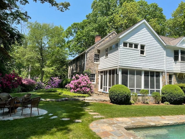 back of property with a sunroom, stone siding, a chimney, a yard, and a patio area