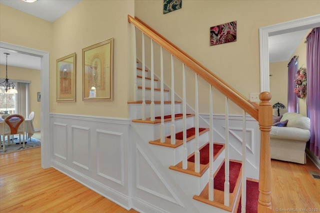 stairs with a wainscoted wall, a decorative wall, wood finished floors, and an inviting chandelier