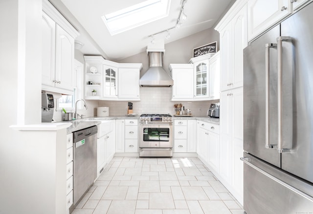 kitchen with vaulted ceiling with skylight, glass insert cabinets, appliances with stainless steel finishes, wall chimney range hood, and backsplash