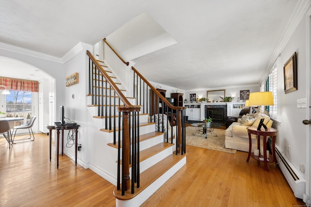 stairway featuring a baseboard heating unit, ornamental molding, wood finished floors, arched walkways, and a glass covered fireplace