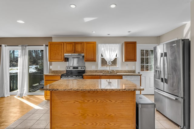 kitchen featuring appliances with stainless steel finishes, brown cabinets, light stone countertops, under cabinet range hood, and a sink