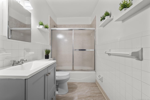 bathroom featuring toilet, shower / bath combination with glass door, vanity, and tile walls