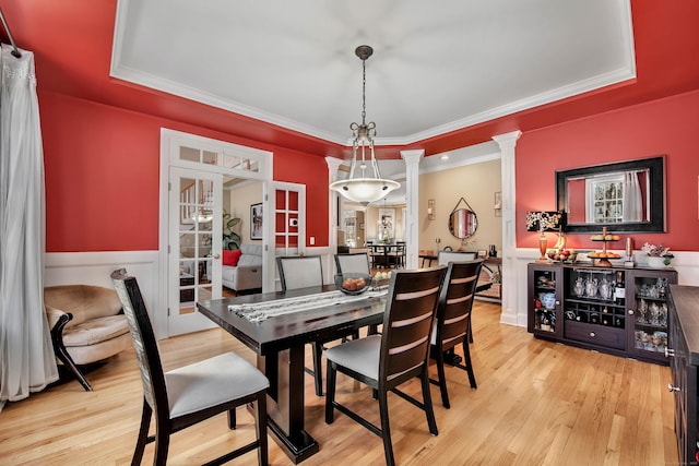 dining area featuring light wood-type flooring, ornate columns, a raised ceiling, and wainscoting