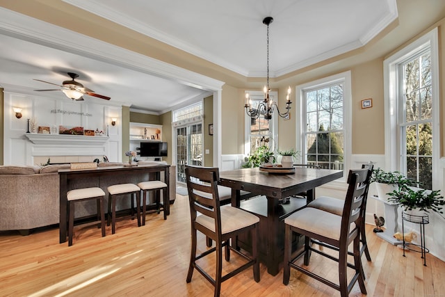 dining area with light wood-style floors, a wainscoted wall, ornamental molding, and ceiling fan with notable chandelier