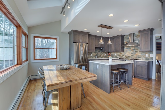 kitchen with lofted ceiling, a baseboard radiator, a kitchen island, appliances with stainless steel finishes, and wall chimney range hood