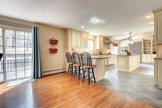 kitchen featuring a center island, baseboard heating, stainless steel appliances, cream cabinetry, and wall chimney range hood