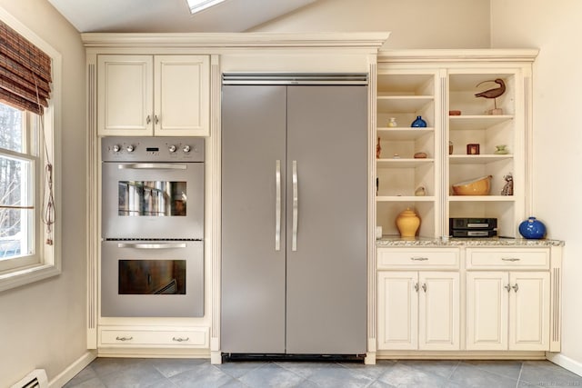 kitchen with built in fridge, cream cabinetry, a baseboard radiator, multiple ovens, and light stone countertops