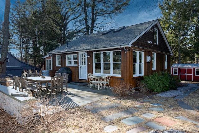 back of property featuring an outdoor structure, outdoor dining area, roof with shingles, and a patio