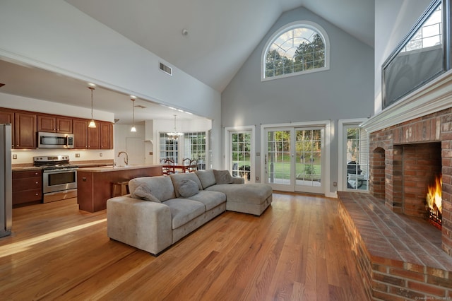 living room featuring high vaulted ceiling, a notable chandelier, wood finished floors, visible vents, and a brick fireplace