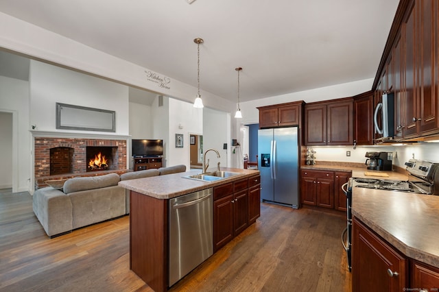 kitchen with a kitchen island with sink, stainless steel appliances, a fireplace, a sink, and dark wood-style floors