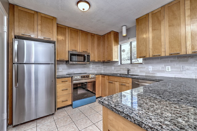 kitchen featuring stainless steel appliances, backsplash, light tile patterned flooring, a sink, and dark stone countertops