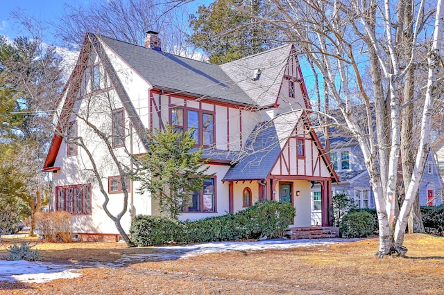 english style home featuring a shingled roof, a chimney, and stucco siding