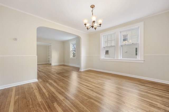 empty room featuring arched walkways, a chandelier, a wainscoted wall, light wood-type flooring, and crown molding