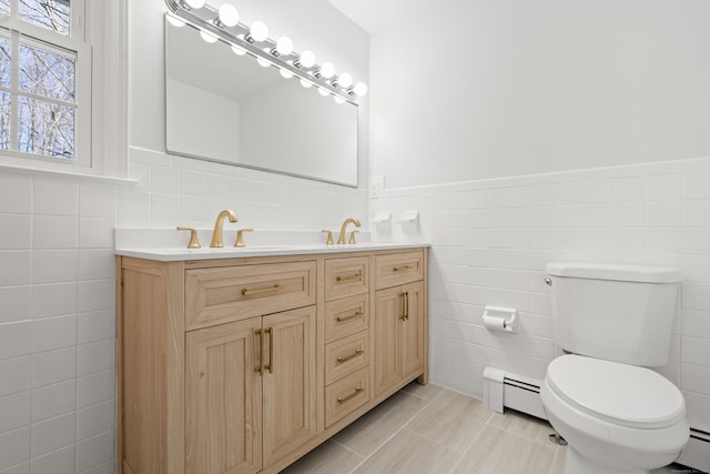 bathroom featuring toilet, double vanity, baseboard heating, tile walls, and a sink