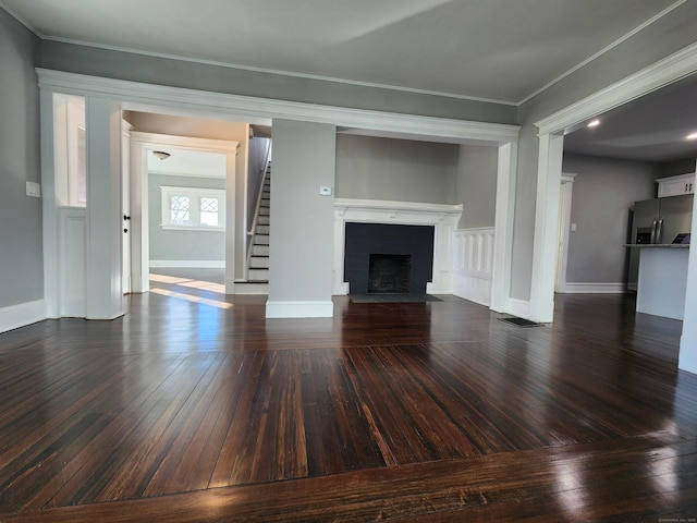 unfurnished living room featuring baseboards, dark wood-style floors, a fireplace with flush hearth, ornamental molding, and stairs