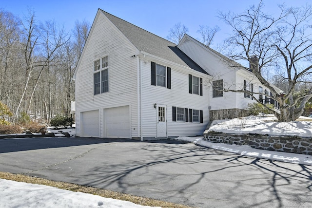 snow covered property with aphalt driveway, an attached garage, and a shingled roof