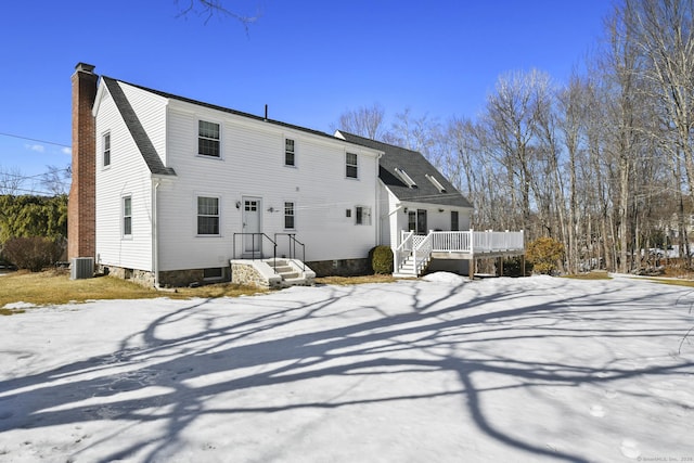 snow covered back of property with a deck, a shingled roof, a chimney, and central AC unit