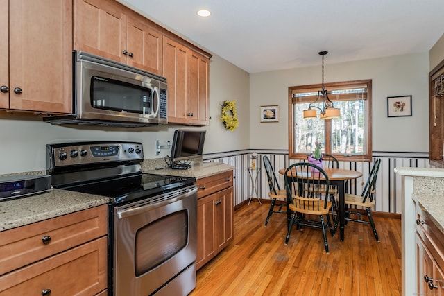 kitchen with a wainscoted wall, pendant lighting, light stone counters, appliances with stainless steel finishes, and light wood-style floors