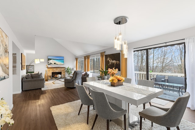 dining area with dark wood-style floors, an inviting chandelier, vaulted ceiling, a stone fireplace, and baseboards