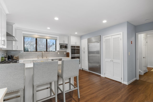 kitchen featuring built in appliances, a peninsula, dark wood-style flooring, a sink, and tasteful backsplash