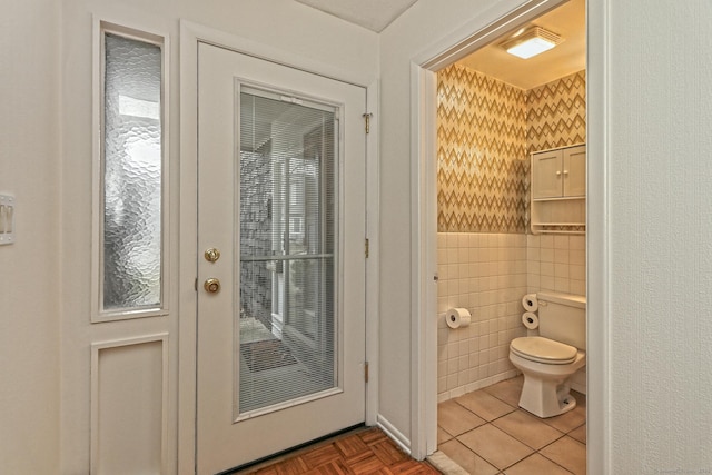 bathroom featuring wallpapered walls, toilet, a wainscoted wall, tile patterned floors, and tile walls