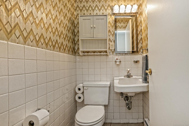 half bath with tile walls, a baseboard radiator, toilet, a sink, and wallpapered walls