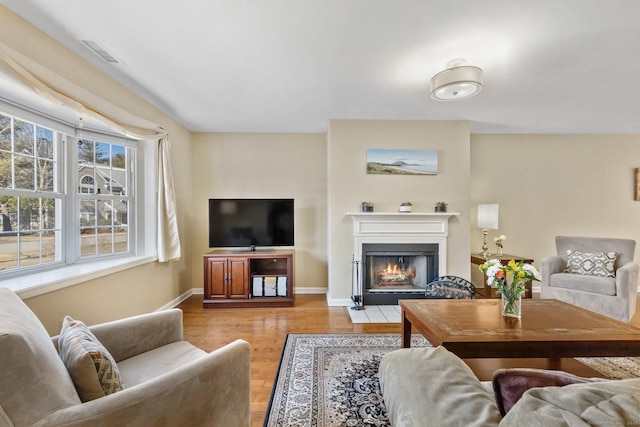 living room featuring baseboards, a fireplace with flush hearth, visible vents, and light wood finished floors