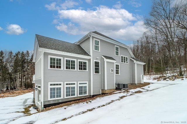 snow covered property featuring cooling unit and roof with shingles