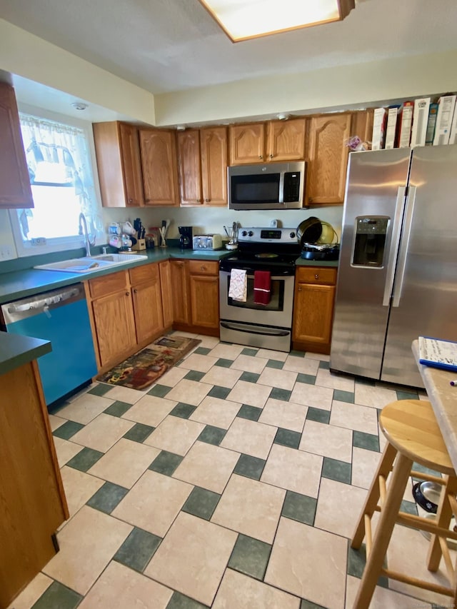 kitchen with light floors, stainless steel appliances, dark countertops, brown cabinetry, and a sink