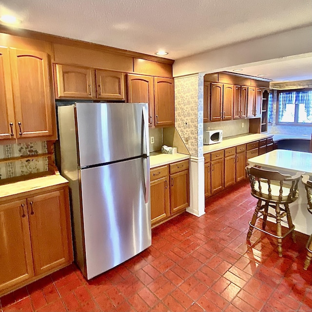 kitchen with white microwave, brick floor, light countertops, freestanding refrigerator, and brown cabinetry