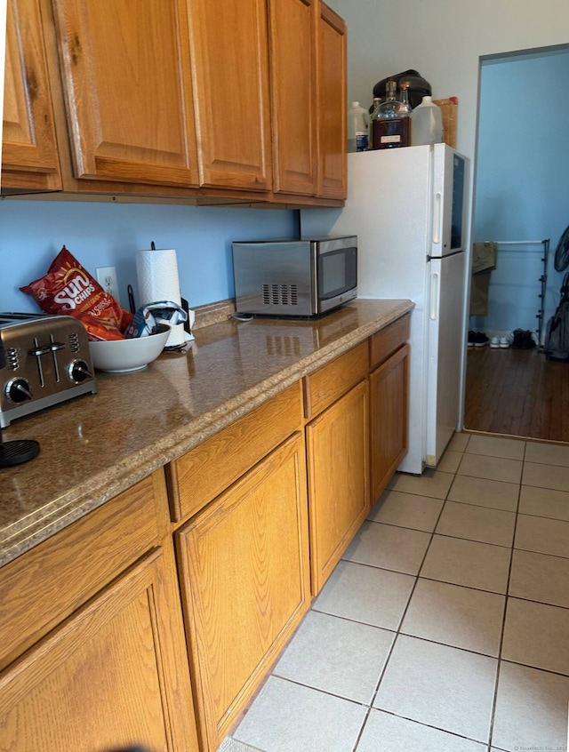 kitchen featuring brown cabinetry, dark stone counters, stainless steel microwave, and light tile patterned flooring