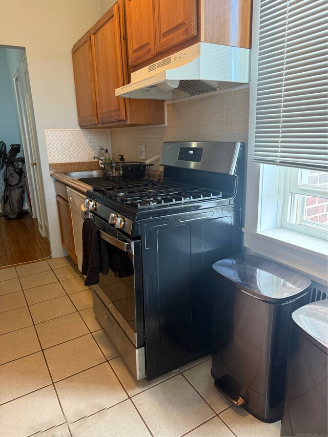 kitchen with light tile patterned floors, under cabinet range hood, brown cabinets, tasteful backsplash, and stainless steel range with gas stovetop